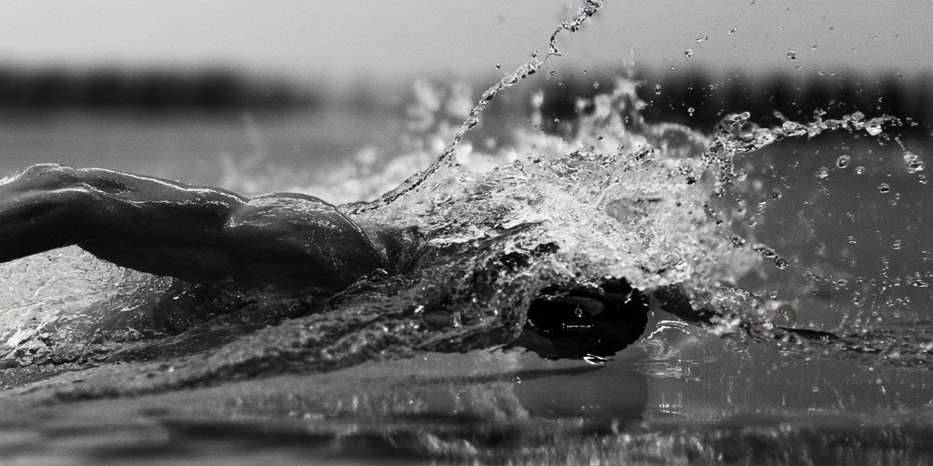 Swimmer's arm recovering above a breaking surface, spray suspended in monochrome